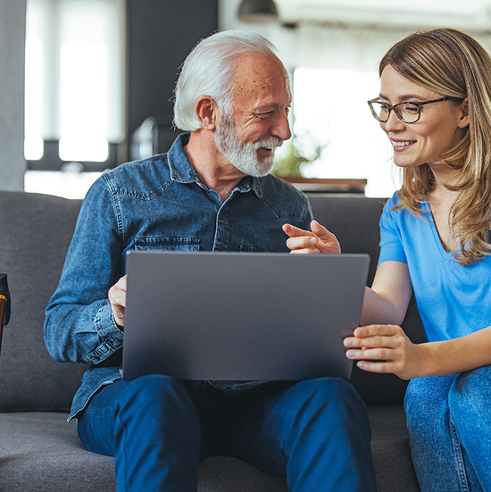 An elderly man with a white beard and a younger woman with glasses are sitting on a couch, smiling while looking at a laptop. The woman points at the screen, both appearing engaged and happy. Theyre in a cozy, well-lit room.