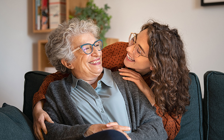 An older woman with gray hair and glasses sits on a couch, smiling warmly. A younger woman with curly hair and glasses embraces her from the side, also smiling. Both appear happy and relaxed in a cozy living room setting.