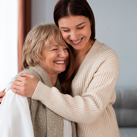 A young woman and an older woman embrace warmly, both smiling with eyes closed. The young woman has long brown hair and wears a beige sweater, while the older woman has short gray hair and wears a white top with a gray scarf. The setting is indoors.